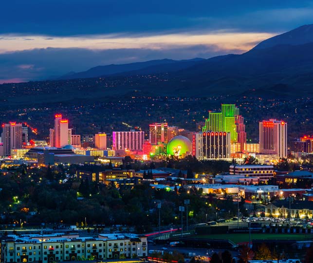 The skyline of Reno, Nevada at night. Bright colors highlight many buildings and the mountains can be seen in the background.