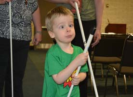 A blind toddler boy stands holding his white cane, two adults with canes stand behind him.